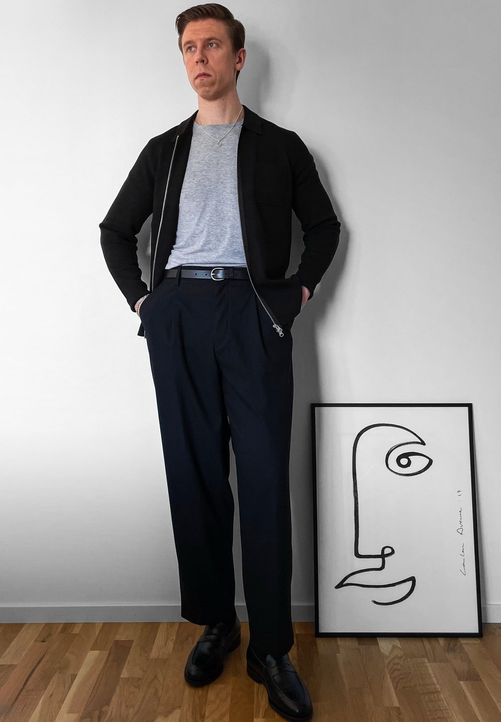 Μαύρο φερμουάρ μπουφάν πάνω από γκρι μπλουζάκι, ναυτικές πτυχές παν trousers, μαύρη ζώνη και μαύρα παπούτσια. Γυρισμένος αφηρημένος σχέδιο γραμμής κοντά.