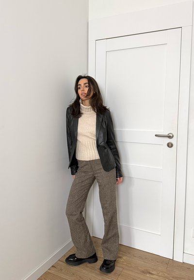 Mujer joven con chaqueta de cuero negra, jersey de cuello alto beige y pantalones marrones, de pie en el interior frente a una puerta y pared blanca.
