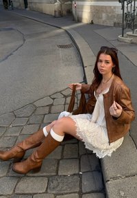 Braune Lederjacke über einem weißen Spitzenkleid, kniehochelige braune Stiefel mit Absatz und eine braune Tasche, sitzend auf Kopfsteinpflaster.