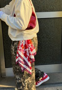 Sac en cuir verni rouge, sweat-shirt blanc, pantalon camouflage et foulard en soie avec motifs rouges et floraux noué à la taille. Baskets rouges visibles.