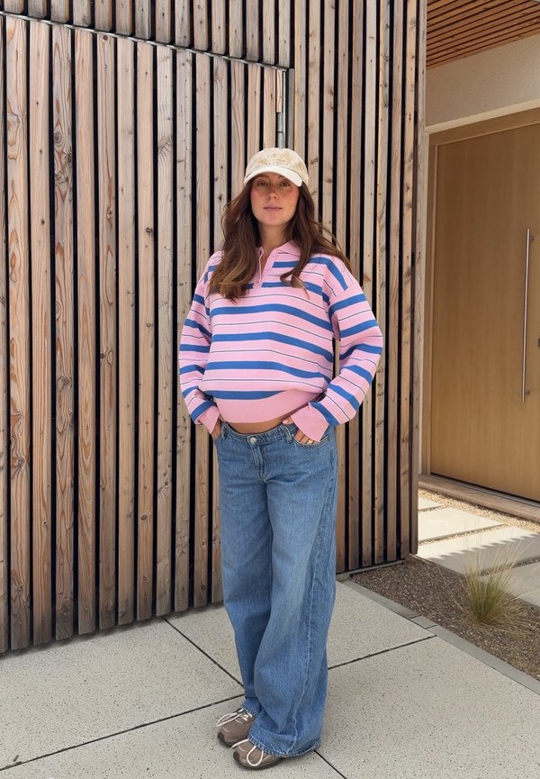 Mujer con suéter de rayas rosas y azules, vaqueros azules holgados, gorra beige y zapatillas, de pie con las manos en los bolsillos junto a una pared de listones de madera y una puerta.