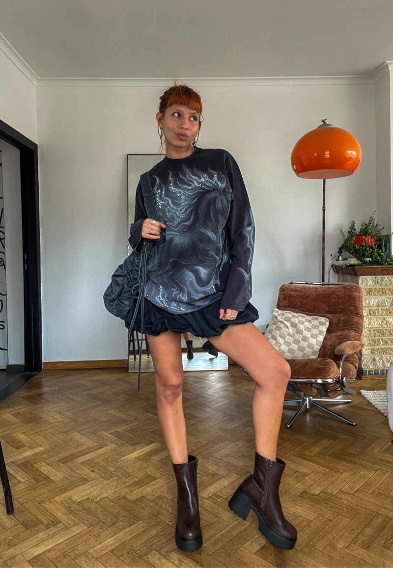 Personne debout à l’intérieur portant un sweat-shirt noir avec un motif de cheval, une jupe noire, des bottes marron épaisses et portant un sac noir avec des plantes et des meubles en arrière-plan.