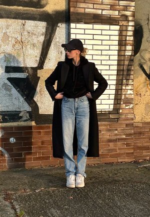 Abrigo largo negro sobre una sudadera negra, jeans de mezclilla azul claro y zapatillas blancas; lleva una gorra negra y gafas; fondo de pared texturizada.