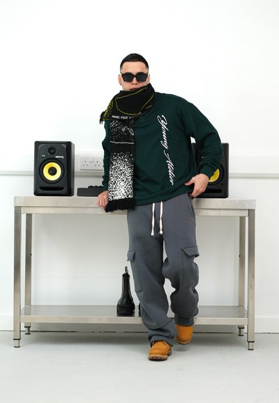 Sudadera verde con texto blanco, pantalones de chándal grises y calzado marrón. Accesorios incluyen una bufanda con patrón negro y amarillo y gafas de sol.