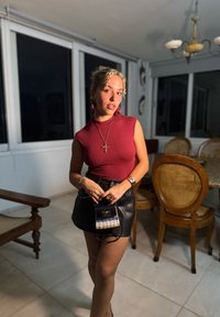 Röd ärmlös topp, svart läderkjol och en liten axelväska med vävt mönster. Lockigt hår och hoopsörhängen. Välbelyst inomhusmiljö.