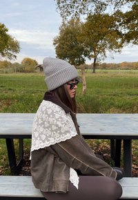 Gray knit beanie, dark jacket with lace accents, and black sunglasses. Sitting at a picnic table outdoors with greenery in background.