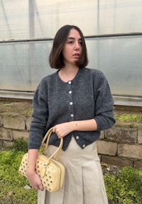 Femme aux cheveux courts et foncés portant un pull gris boutonné et une jupe beige, tenant un sac à main jaune avec des œillets métalliques en plein air.