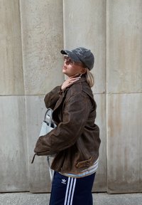 Καφέ δερμάτινο μπουφάν, γκρι καπέλο με κεντήματα, ανοιχτόχρωμα γυαλιά ηλίου, μπλε και λευκή ριγέ πουκάμισο, μαύρο παντελόνι φόρμας με άσπρες ρίγες, ασημένια τσάντα.