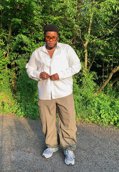 Camisa blanca de botones con dos bolsillos en el pecho, combinada con pantalones cargo marrón claro y zapatillas deportivas grises. Fondo de follaje verde.