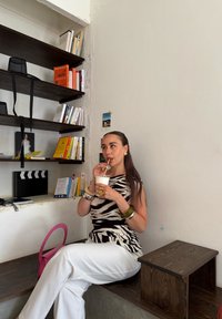 Zebra-gestreepte top, brede witte broek, nippend uit een kopje. Houten plank op de achtergrond met boeken. Roze handtas en metalen armbanden.