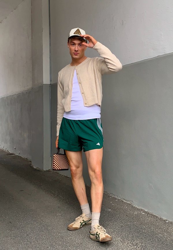 Beiger Strickcardigan über einem weißen Hemd, grüne Sportshorts, weiß-grüne Sneaker und eine karierte Handtasche. Schirmmütze nach vorne getragen.