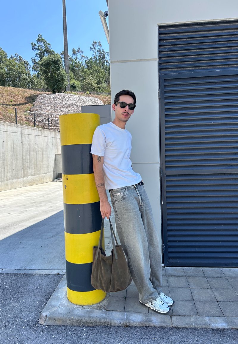 Homme portant des lunettes de soleil, un t-shirt blanc et un jean, appuyé contre un poteau rayé jaune et noir, tenant un sac marron à l'extérieur.