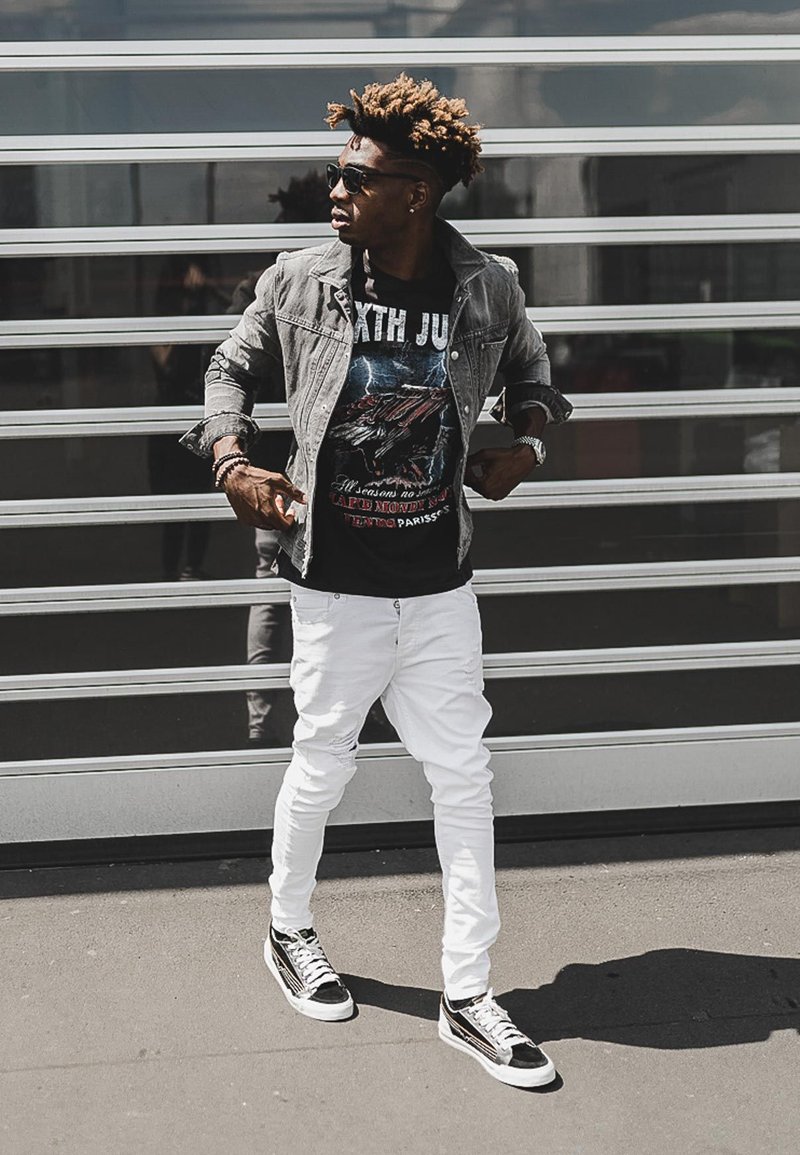 Denim jacket, graphic black t-shirt featuring a bird design, white ripped jeans, and black sneakers with white accents. Sunglasses complete the outfit.