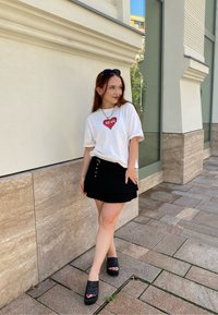 Bijela majica s grafikom crvenog srca, uparena s crnom plisiranom suknjom. Crne platforme upotpunjuju cijeli outfit. Moderan urbani ambijent.