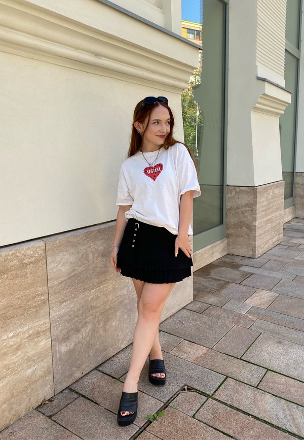 Biela tričko s červenou grafikou srdca, v kombinácii s čiernou plisé sukňou. Čierne platformové topánky dokončujú outfit. Štýlové mestské prostredie.