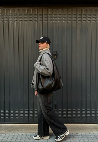 Gran bolso tote de cuero negro grande con una textura suave y lisa; combinado con una chaqueta gris de gran tamaño y jeans oscuros. Fondo de ladrillo gris.