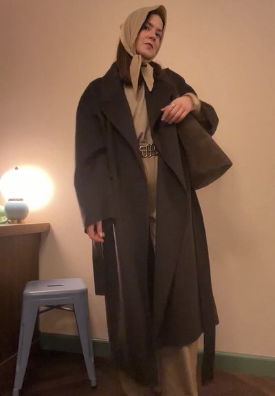 Mujer con pañuelo beige en la cabeza, abrigo y conjunto a juego, de pie en el interior cerca de una lámpara sobre una mesa y un taburete de metal.