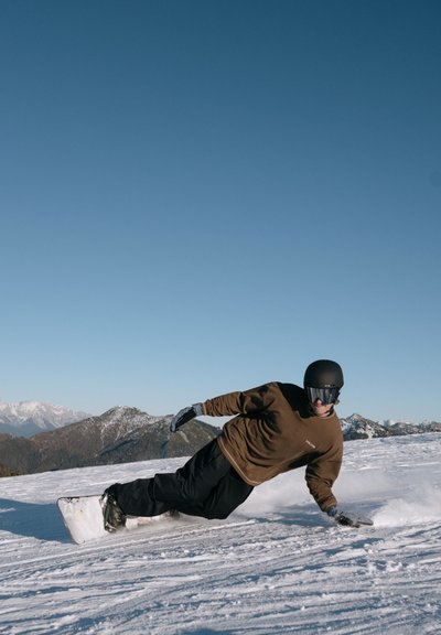 Snowboarder con camiseta marrón de manga larga y pantalones negros, llevando un casco negro y gafas, esculpiendo en la nieve con montañas al fondo.