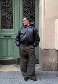 Γυναίκα με μαλλιά πιασμένα πίσω στέκεται με τα χέρια στις τσέπες, φορώντας μαύρο δερμάτινο μπουφάν και φαρδιά γκρι παντελόνια, δίπλα σε πράσινη περίτεχνη πόρτα.