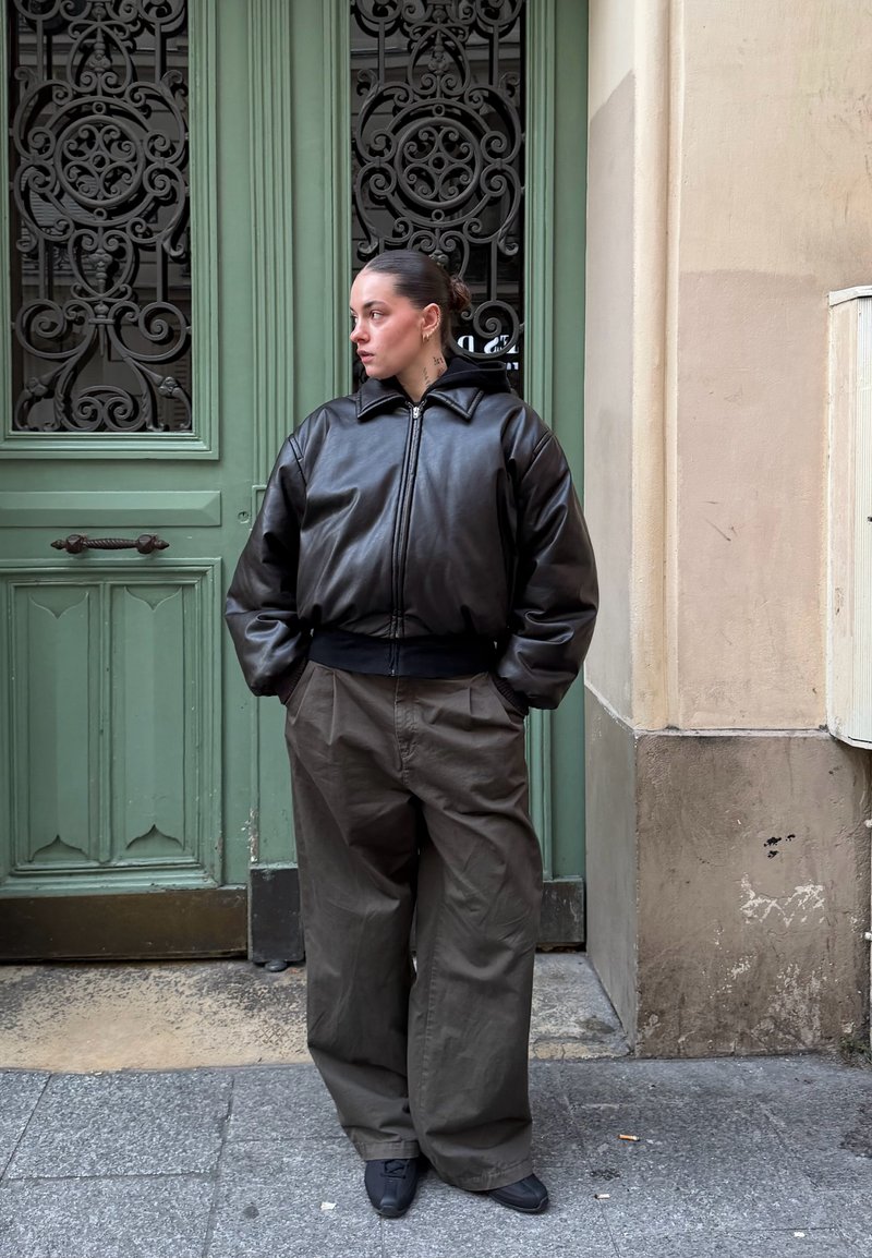 Femme aux cheveux attachés, les mains dans les poches, portant une veste en cuir noire et un pantalon gris surdimensionné, devant une porte verte ornée.