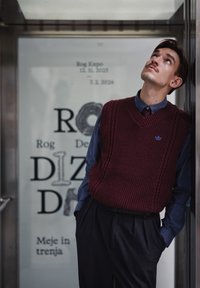 Gilet en tricot marron avec un col en V, chemise bleue boutonnée en dessous, et pantalon foncé. Accent de logo bleu sur le gilet. Cadre urbain moderne.