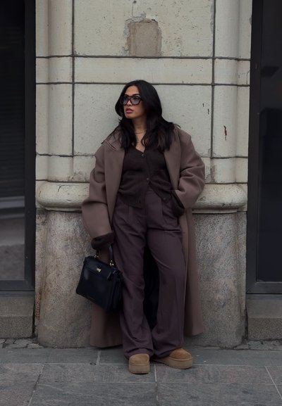 Abrigo marrón oversize, suéter marrón oscuro, pantalones marrón de pierna ancha, zapatos de plataforma color beige y un bolso negro con acentos dorados. Fondo urbano.