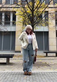 Λευκή γούνα πάνω από λευκή μπλούζα, ανοιχτό μπλε χαλαρές τζιν, μπεζ πλατφόρμες και κρέμα καπέλο, στέκεται σε εξωτερικό χώρο.