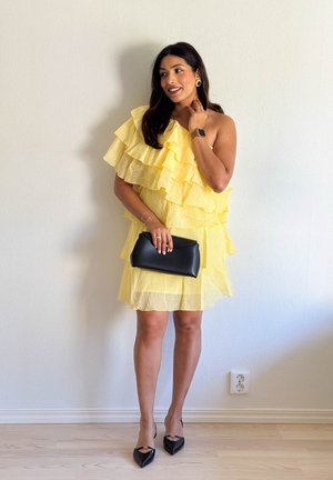 Vestido amarillo de un solo hombro con volantes en capas, fabricado con un tejido texturizado, combinado con un clutch negro y tacones negros de punta. Accesorios mínimos.