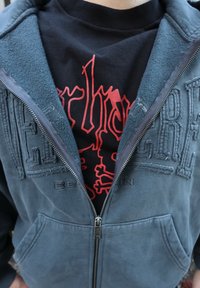 Sweat à capuche zippé gris en tissu doux, avec un logo texturé, un col doublé en polaire et des poches avant. T-shirt noir en dessous avec impression rouge.