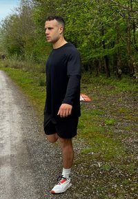 Melna garā piedurknēm krekls, melni šorti, baltas un melnas apavus ar sarkaniem akcentiem, dodas pa gružaino ceļu, ko ierāmē zaļums.