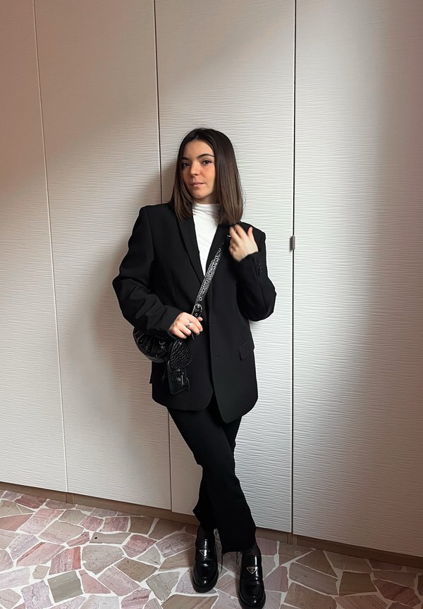 Junge Frau in schwarzem Blazer und Hose mit weißem Oberteil, hält eine schwarze Tasche und steht vor einer hell strukturierten Wand auf gemustertem Boden.