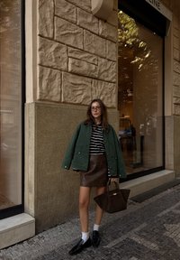 Vrouw in een groene jas over een zwart-wit gestreepte shirt, bruine leren rok, witte sokken, zwarte loafers en met een grote bruine handtas in haar handen.