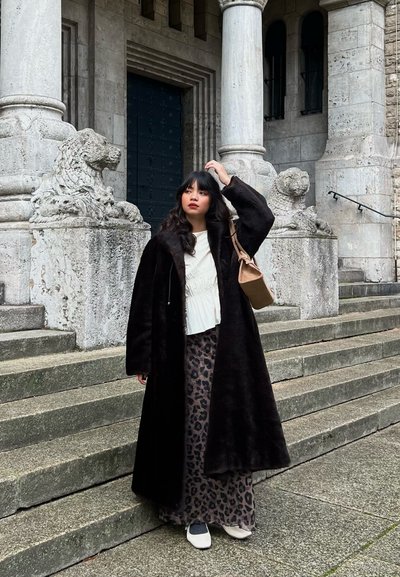 Abrigo de piel sintética negra sobre una blusa blanca texturizada y una falda larga con estampado de leopardo, combinado con zapatos blancos y grises, de pie sobre escalones de piedra.