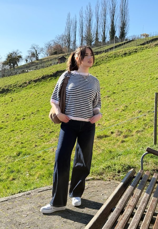 Young woman wearing a striped jumper and dark trousers stands on a path next to a bench, with a green grassy hill and trees in the background.