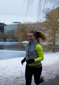 Femme en jogging portant un haut à manches longues jaune vif et un gilet gris, courant sur un chemin enneigé près de l'eau avec des bâtiments et des arbres nus en arrière-plan, souriante.
