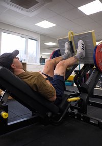 Máquina de prensa de piernas con un usuario empujando una plataforma con peso. El usuario lleva una camiseta marrón y pantalones cortos azul marino, usando zapatos deportivos blancos. Ambiente de gimnasio interior.