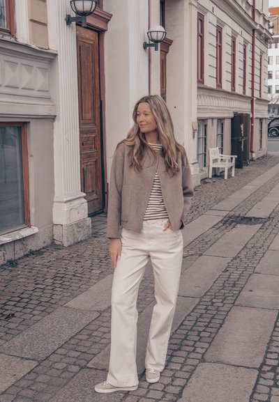 Mujer con pelo largo, chaqueta beige y pantalones blancos, de pie en una calle empedrada cerca de edificios históricos con puertas de madera.