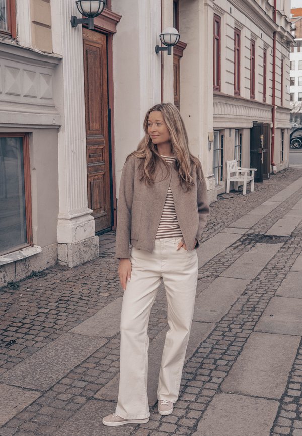 Žena s dugom kosom u bež jakni i bijelim hlačama stoji na kaldrmnoj ulici blizu povijesnih zgrada s drvenim vratima.