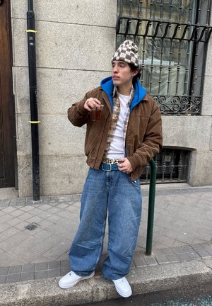 Joven con pantalones vaqueros extragrandes, chaqueta marrón, sudadera azul con capucha y gorra a cuadros, sosteniendo una bebida mientras está de pie en una acera de la ciudad cerca de un poste de metal.
