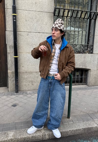 Joven con pantalones vaqueros extragrandes, chaqueta marrón, sudadera azul con capucha y gorra a cuadros, sosteniendo una bebida mientras está de pie en una acera de la ciudad cerca de un poste de metal.