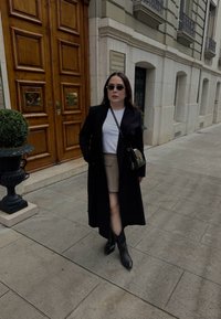 Czarny długi płaszcz na białej koszulce, beżowej spódnicy i czarnych butach na szpilce. Dodatki to mała czarna torebka na ramię i okulary przeciwsłoneczne.