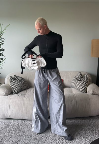 Top negro ajustado de manga larga, pantalones deportivos grises de gran tamaño y un bolso blanco con detalles negros. Fondo neutro y sofá suave.