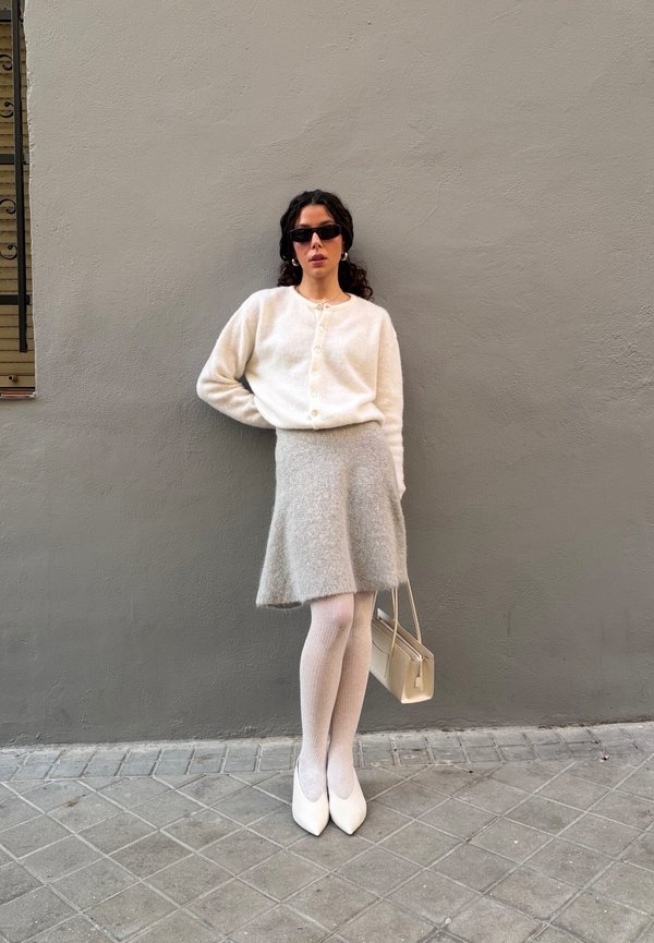 Cardigan blanc en tricot avec boutons, jupe évasée grise, collants blancs côtelés, escarpins blancs pointus, et sac à main beige. Cheveux bouclés et lunettes de soleil.