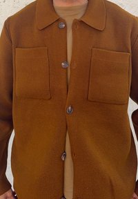 Brown textured button-up jacket featuring two front pockets, a collar, and five buttons in a contrasting colour. Underneath is a tan shirt.