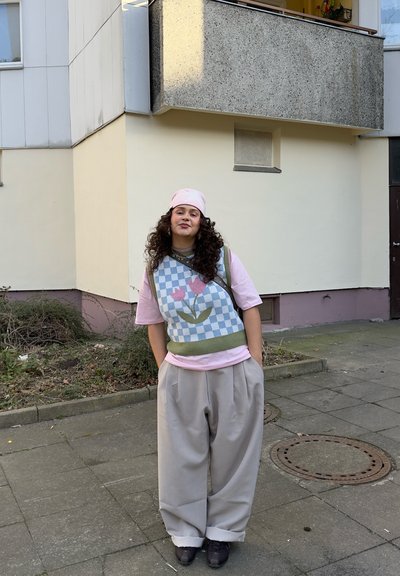 Persona joven con cabello rizado que lleva un gorro rosa, camisa rosa, chaleco de punto con flores y pantalones grises sueltos, está de pie en una acera junto a un edificio.
