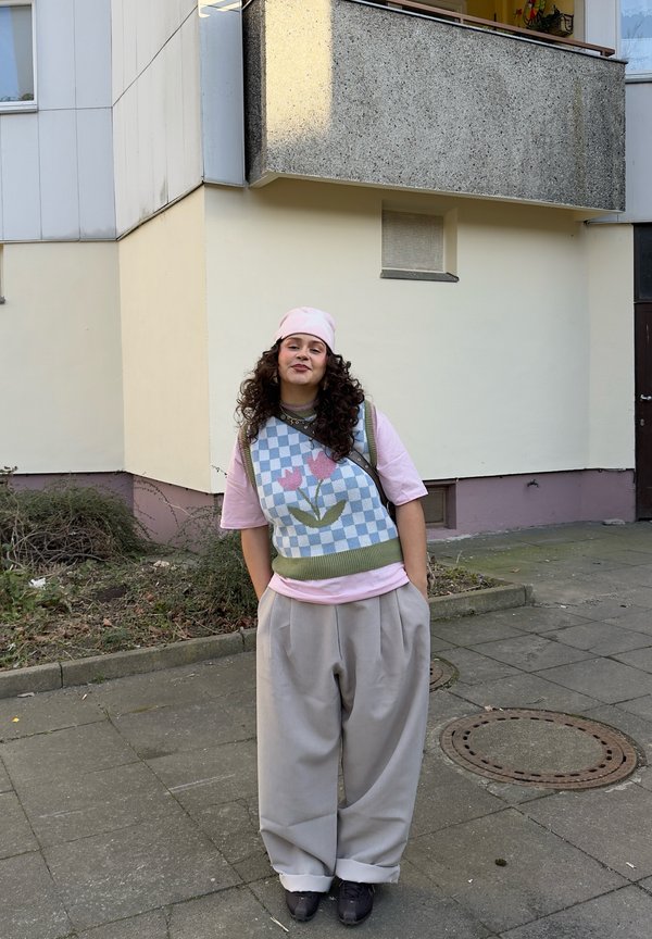 Mladá osoba s kučeravými vlasmi, ktorá má na sebe ružovú čiapku, ružovú košeľu, kvetovaný sveter bez rukávov a voľné sivé nohavice, stojí na chodníku pri budove.