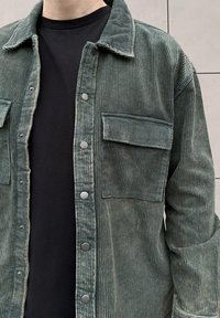 Chemise en velours côtelé vert avec des rayures horizontales, des boutons pression et une poche poitrine. Portée par-dessus un T-shirt noir.