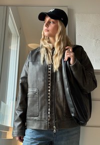 Casaco de pele sintética castanho com fecho de correr e bolsos inclinados, sobre uma blusa às riscas em preto e branco. Boné preto e jeans de ganga.