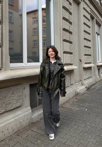 Lesklá tmavě zelená kožená bunda, vzorovaná košile, šedé kalhoty s širokými nohavicemi a stříbrné tenisky. Chodí kolem kamenné budovy s velkými okny.