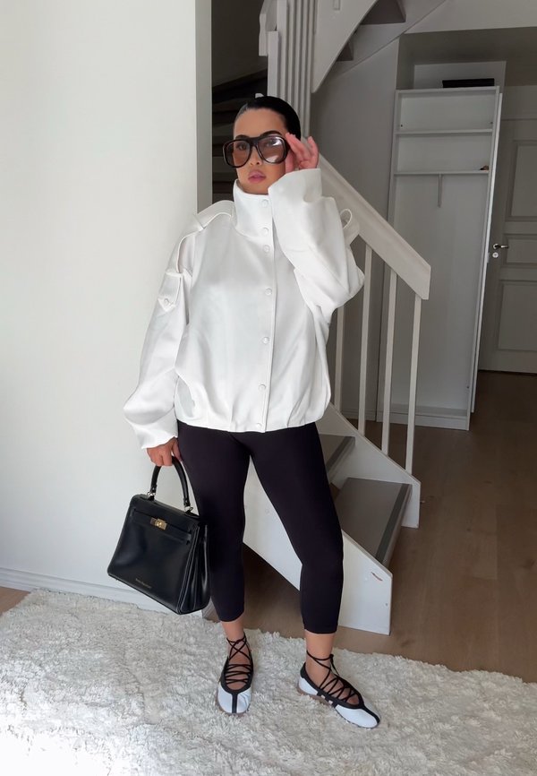 Femme portant une veste blanche à col montant, des leggings noirs courts, de grandes lunettes de soleil, et tenant un sac à main noir près d'un escalier intérieur.